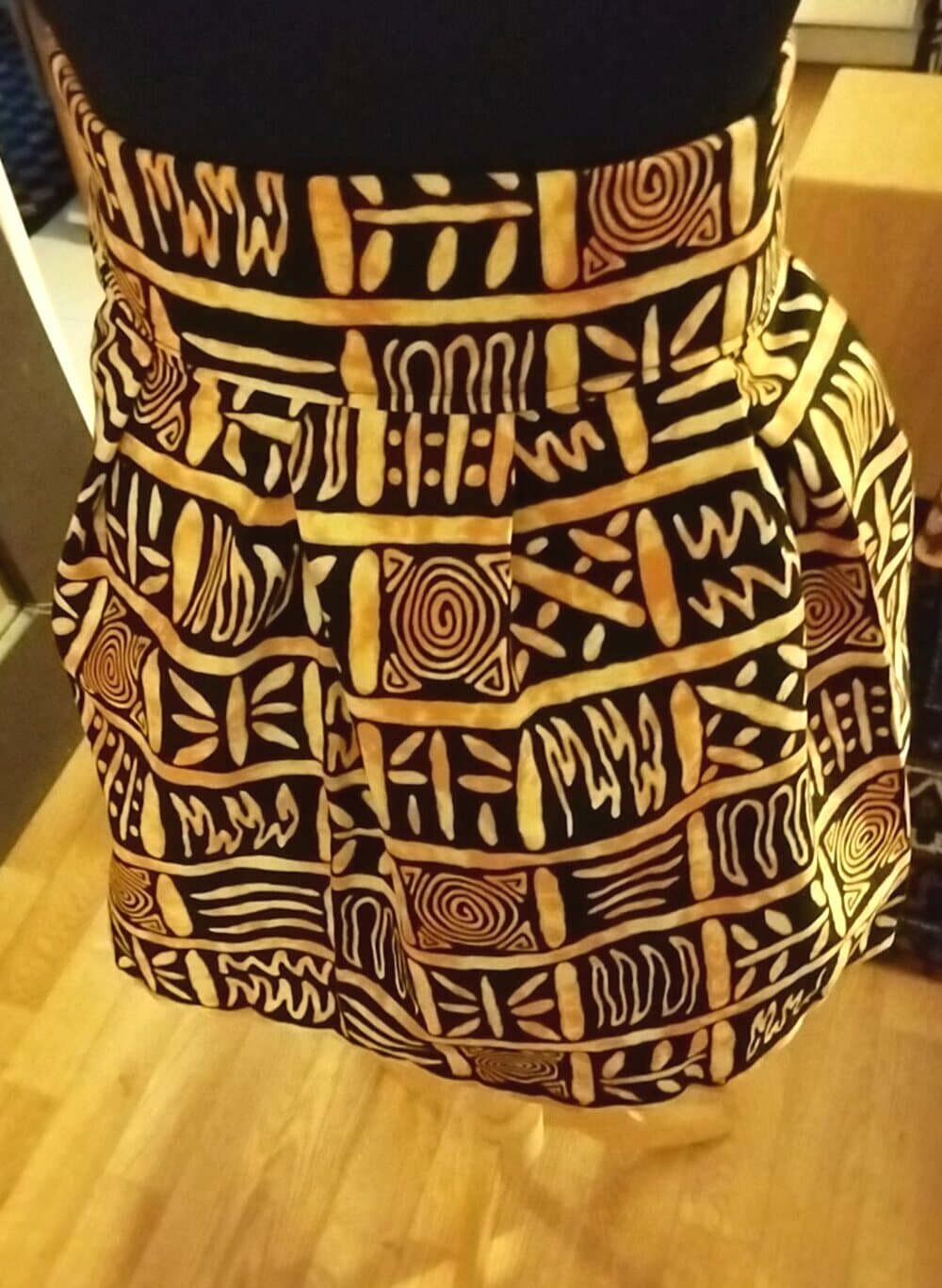 La jupe courte plissée Sable portée : les plis tombent depuis une ceinture large, motif ocre et jaune sur fond noir.