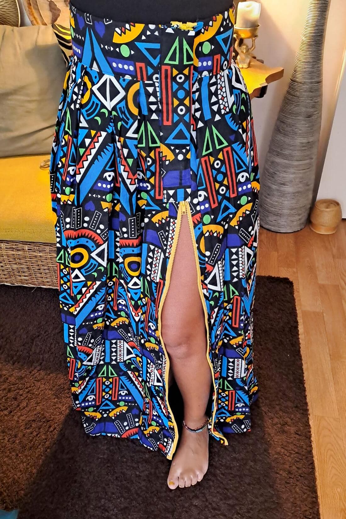 Jupe longue en Wax à motifs géométriques bleus, rouges, jaunes, verts et blancs sur fond noir, portée, la fente devant fermée par une glissière jaune.
