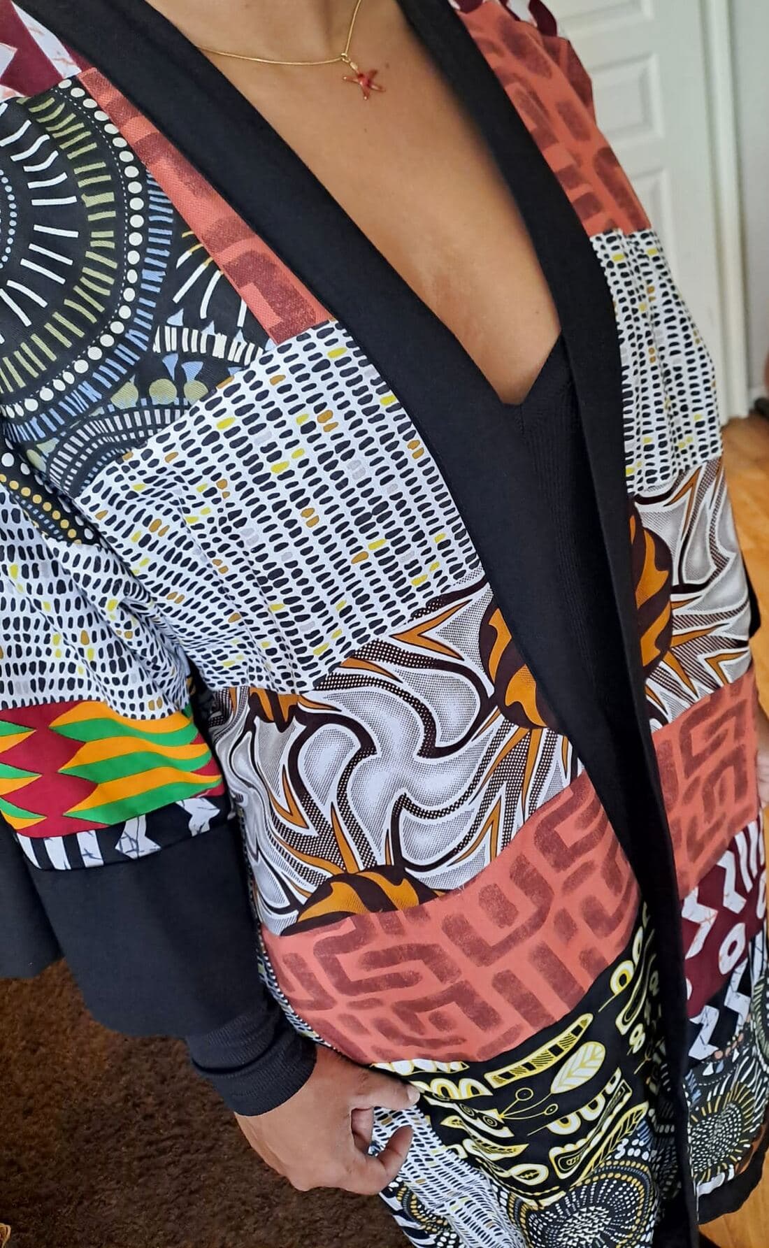 Kimono long à pans ouverts, en patchwork de Wax noir, blanc, ocre et terracotta, porté ouvert sur un haut noir, col, poignets et ourlet bordés de jersey noir.